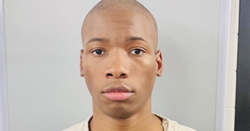 Young man with a shaved head and neutral expression, wearing a light-colored shirt, standing against a plain background.