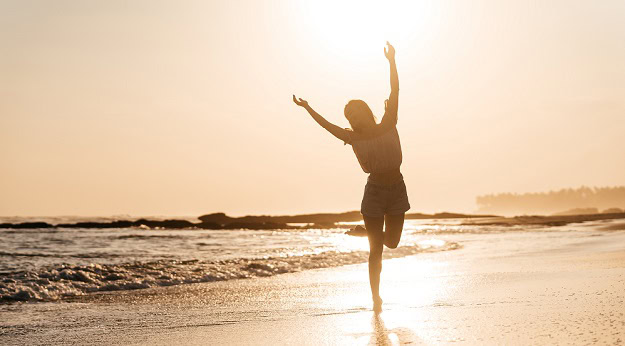 110 Uplifting Quotes to Inspire and Motivate You Right Now A happy woman dancing on the beach by the waters edge.