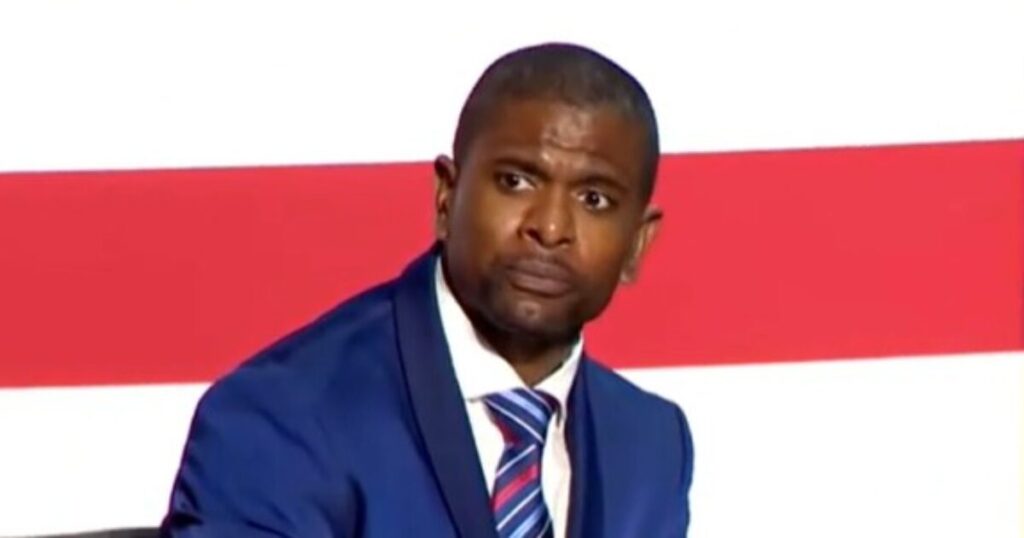 “Disgusting and Sad” – Ex-Vikings Star Jack Brewer Goes Off on WNBA’s Cheryl Reeves for Promoting Child Genital Mutilation with “Trans Kids Belong” Shirt Man in a blue suit with a concerned expression sits against a backdrop of red and white stripes, conveying a serious moment during a discussion or event.