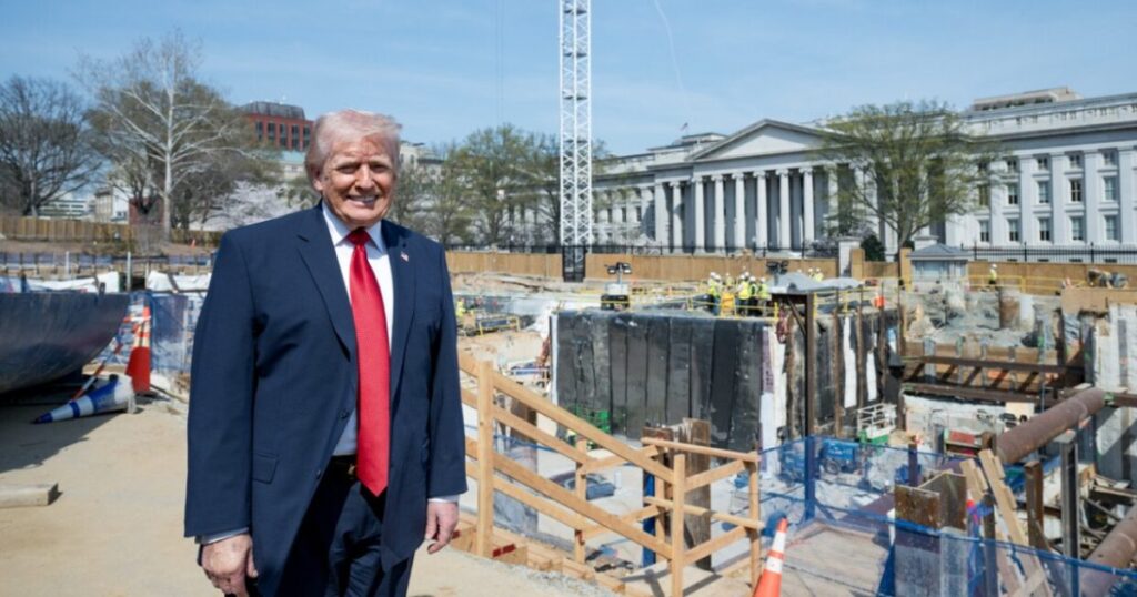 Former President Donald Trump stands in front of construction site near the U.S. Treasury, with workers and equipment visible in the background.