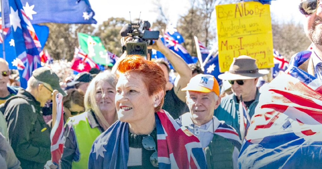 Crowd of protesters holding Australian flags at a rally, featuring a woman in the foreground with bright red hair and a flag-themed outfit.