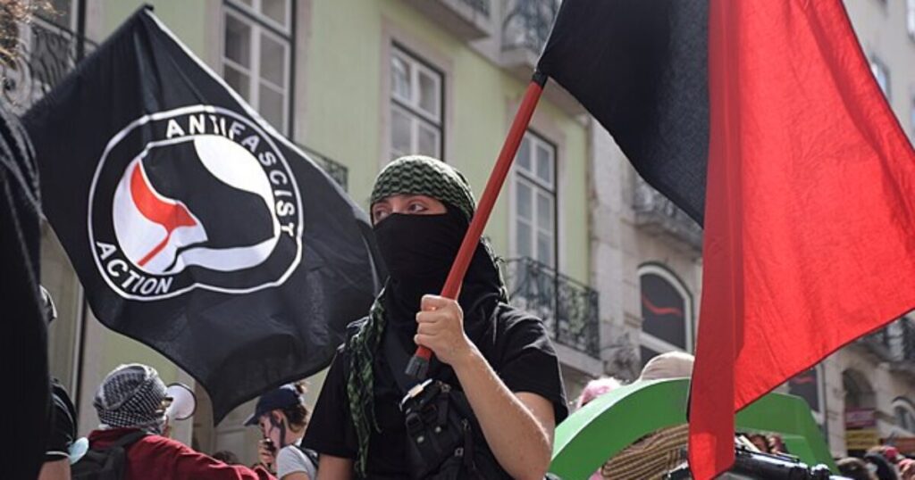 Trump State Department Designates Antifa Linked Group as Global Terrorists * The Gateway Pundit * by Mike LaChance Person in a black mask and green headscarf holding a red flag at an anti-fascist rally with banners in the background.