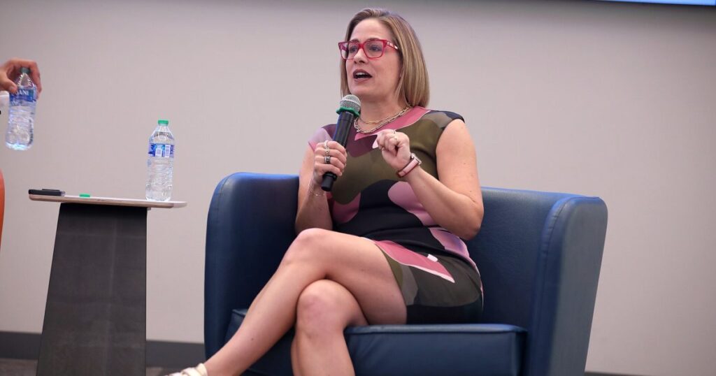 Judge Lets ‘Homewrecker’ Lawsuit Against Kyrsten Sinema Proceed After Finding Her Testimony Not Credible A woman in a colorful dress speaks into a microphone while seated on a blue chair during a discussion event.