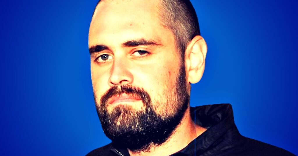 Close-up portrait of a man with a beard and shaved head, set against a blue background, expressing a serious demeanor.