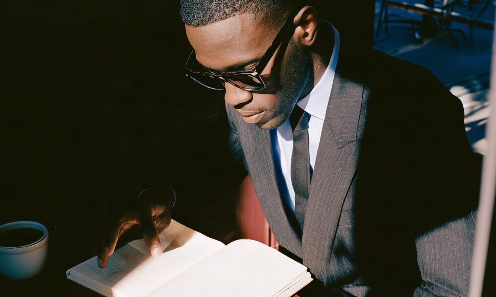 7 Quiet Signs You’re Making Far More Progress Than You Think a black male founder in his mid-30s working early in the morning at a minimalist dark walnut desk to improve his life and ahcive more success