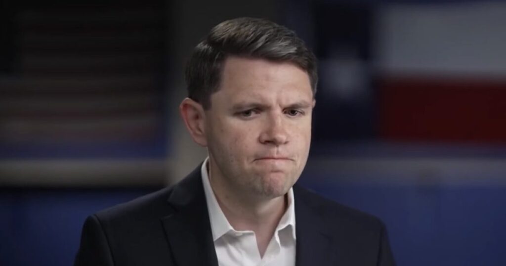Man in a suit with a concerned expression, sitting in front of a blurred Texas flag background.
